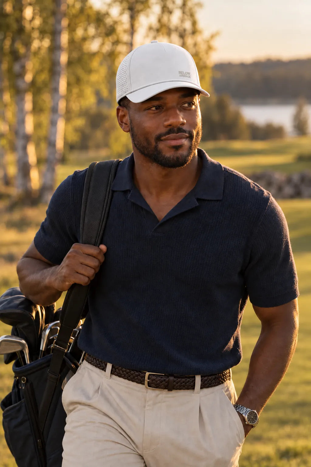 Man in a navy knitted golf polo with his golf bag on the fairway in evening sun, white cap with small embroidered text